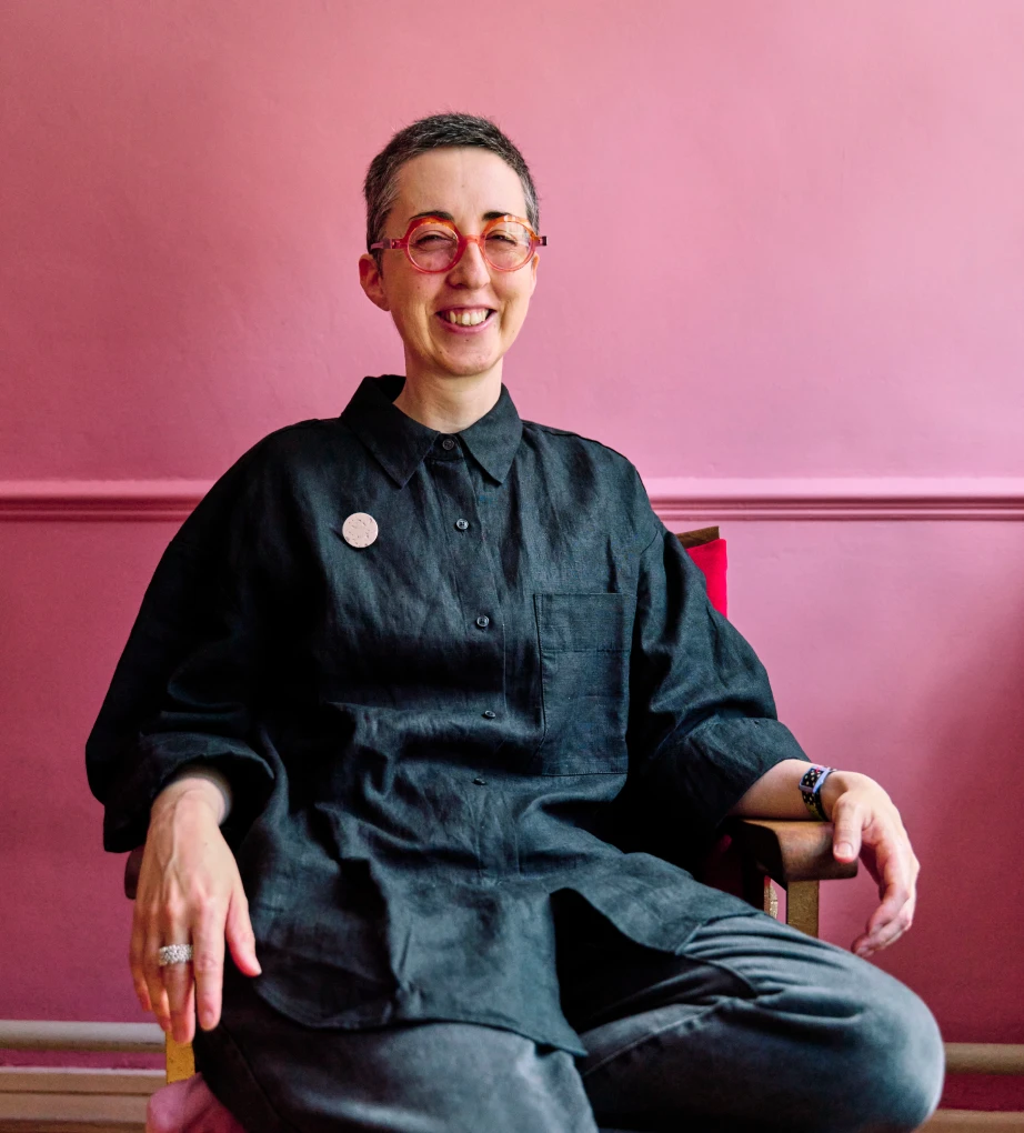 Hannah smiling at the camera, wearing a black shirt, red glasses, against a milkshake pink wall.