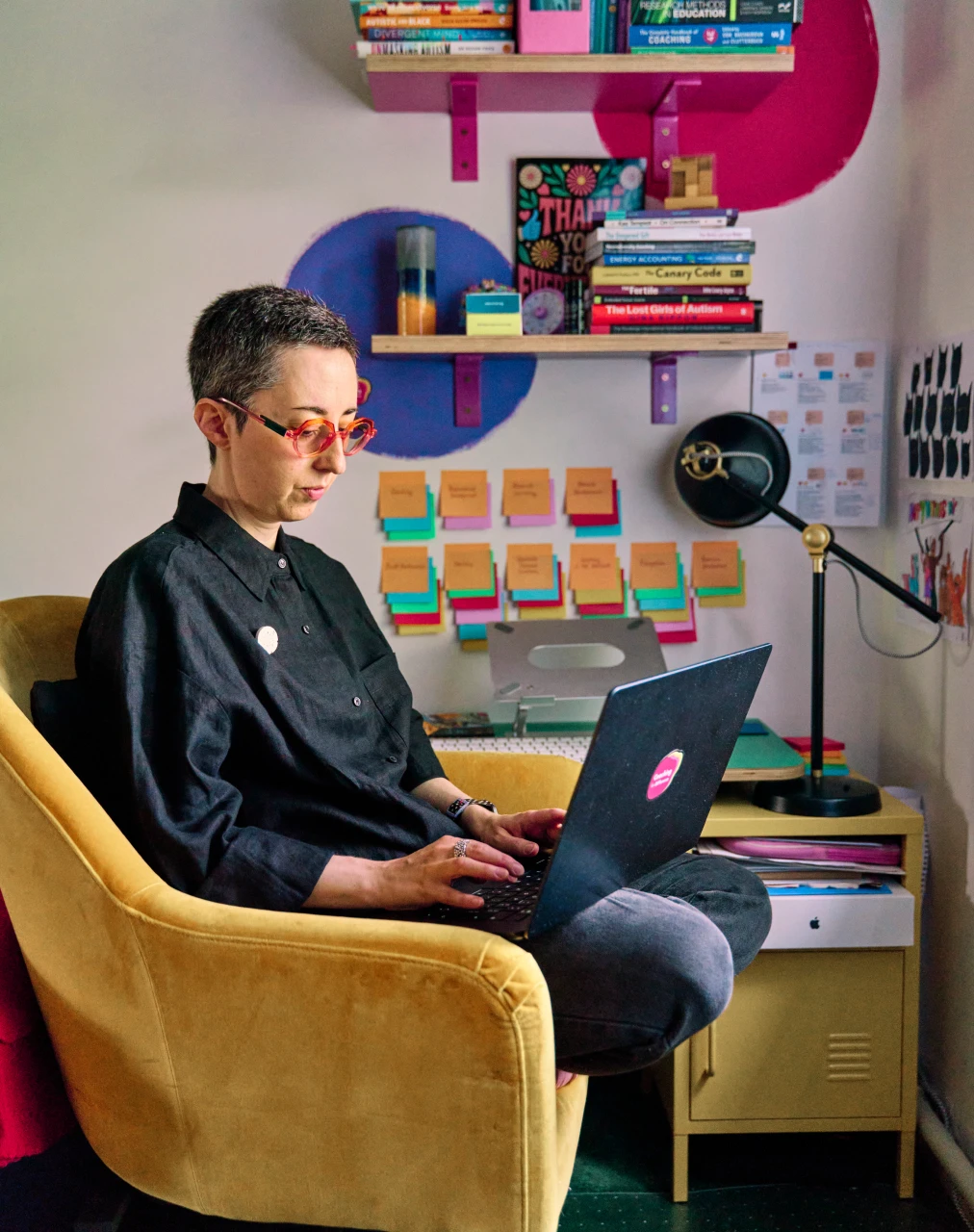 Hannah working in her creative colourful office, sitting in a yellow chair, with laptop.